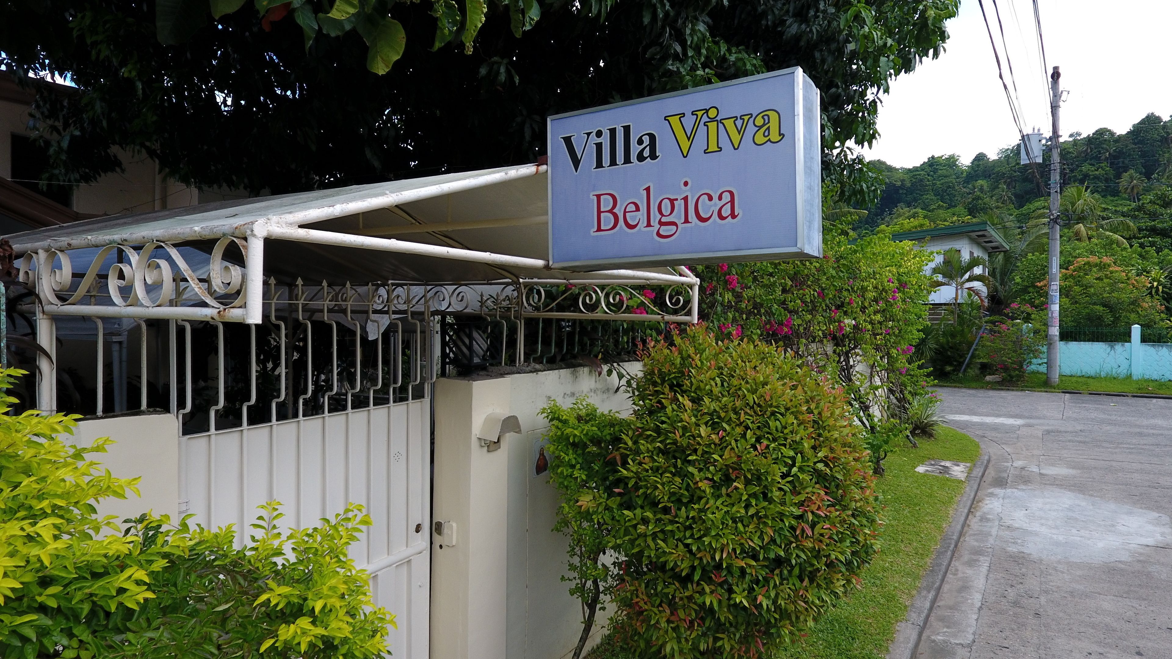 A sign for Villa Viva Belgica, a gated property with a metal gate and a covered walkway, surrounded by lush greenery and a residential area.