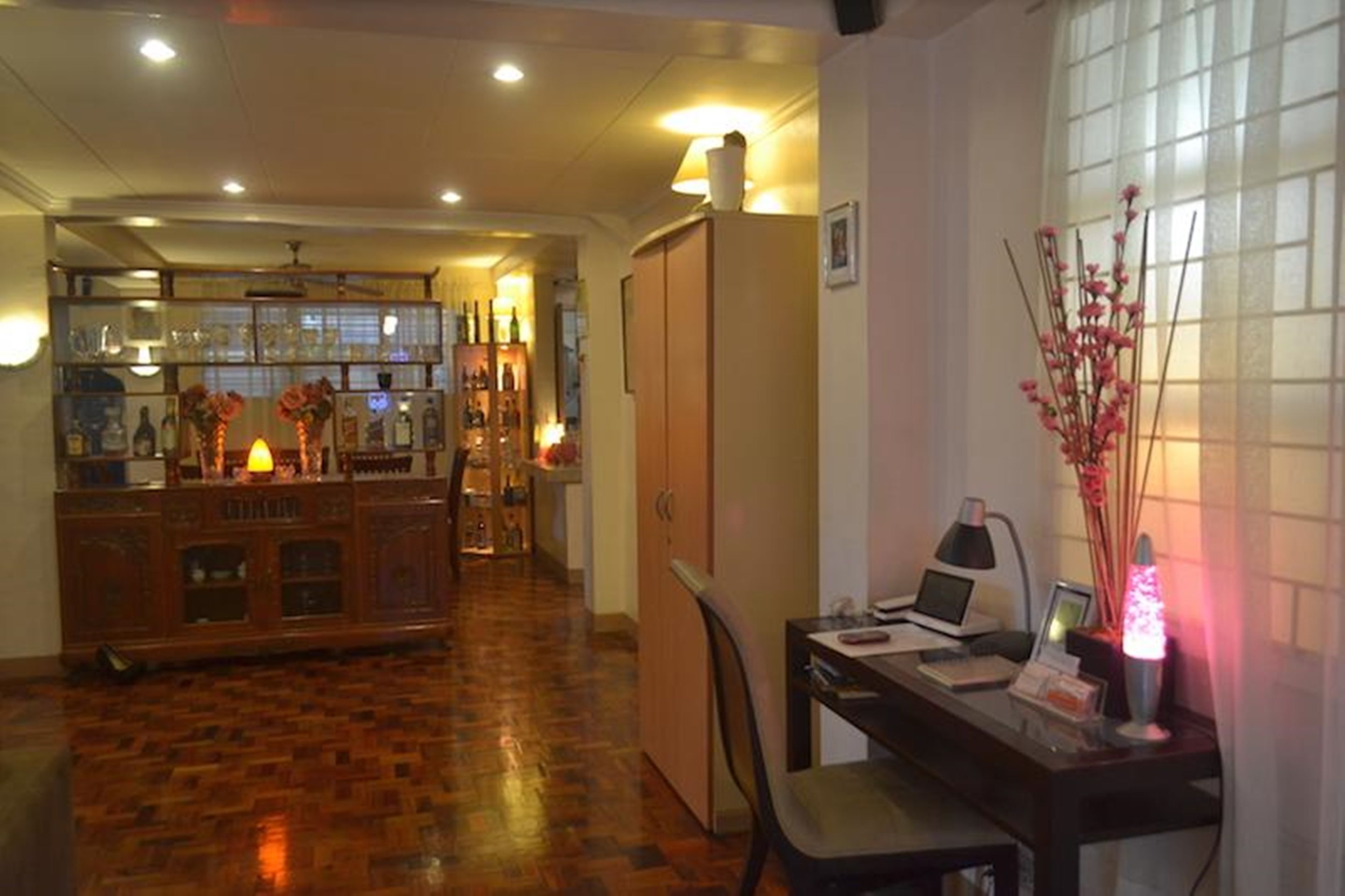 A room with a wooden floor, a desk with a computer, a chair, a lamp, and a bar area with bottles and a shelf.