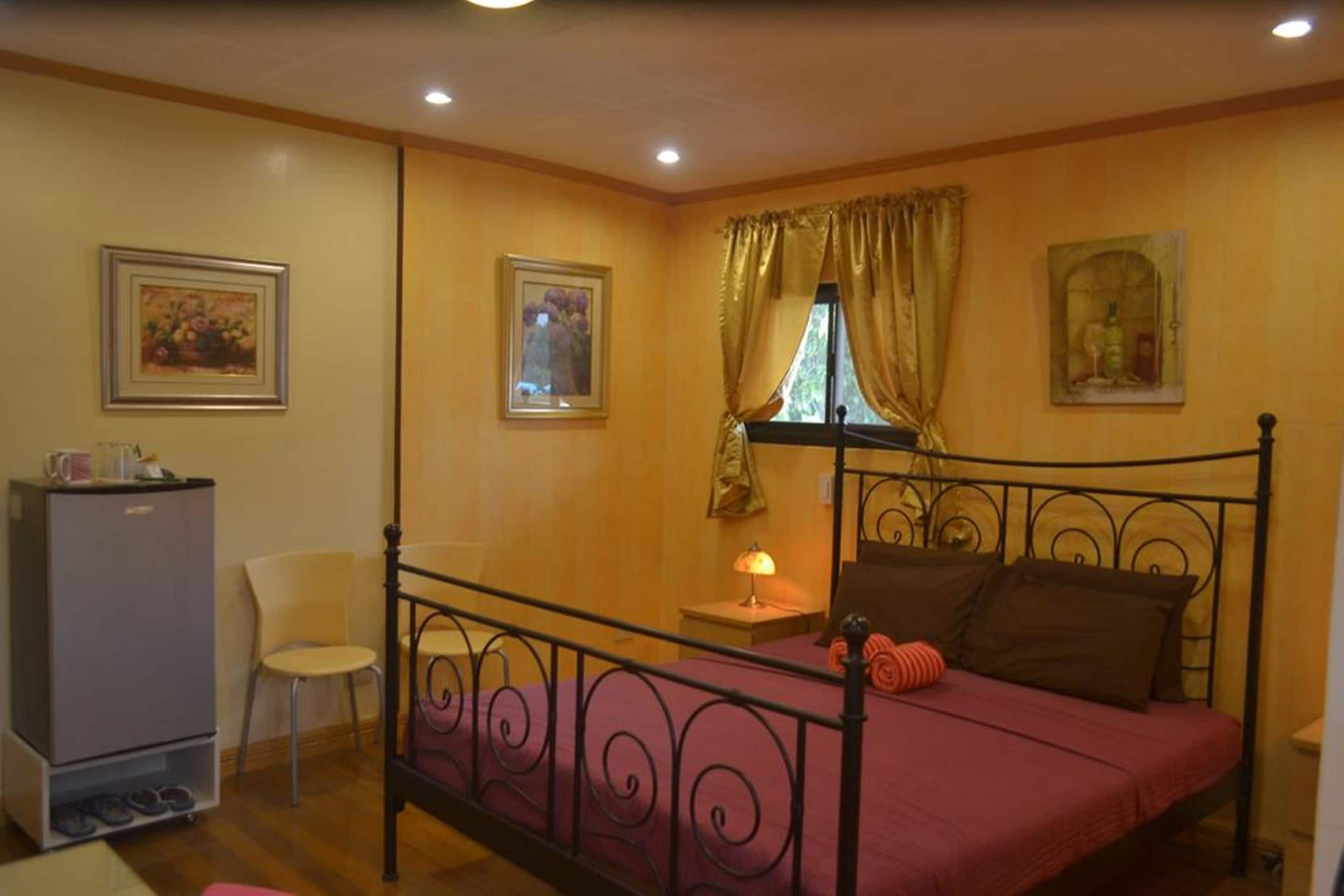A bedroom with a metal bed frame, a single bed with a red bedspread, a small bedside table with a lamp, a chair, and a refrigerator.