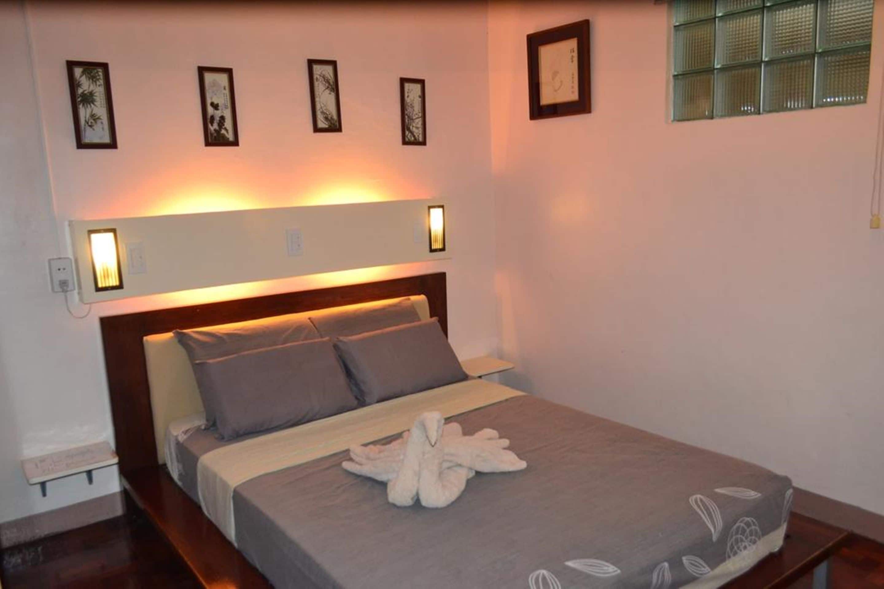 Un lit bien fait, avec une couverture grise et des oreillers, une serviette blanche posée sur le lit, et des lampes fixées au mur au-dessus du lit.