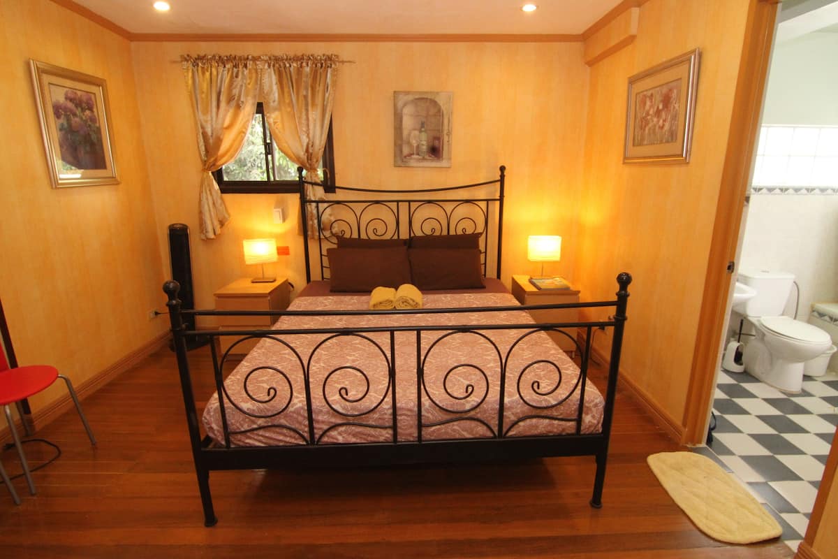 Un dormitorio con estructura de cama metálica, dos lámparas de noche, un ventanal con cortinas, suelo de madera y un piso a cuadros en el baño.