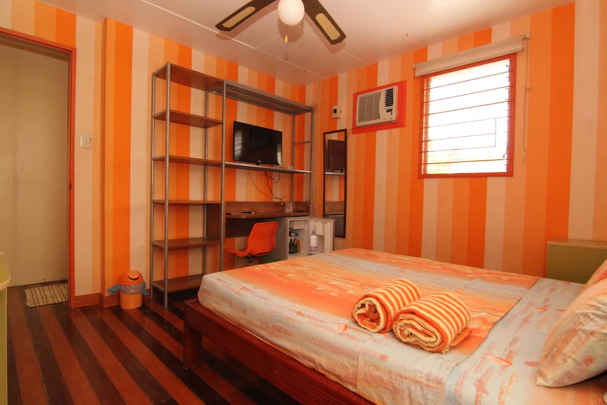 Una habitación con paredes anaranjadas y blancas a rayas, una cama con manta a rayas, un televisor en un estante y una ventana con persianas.
