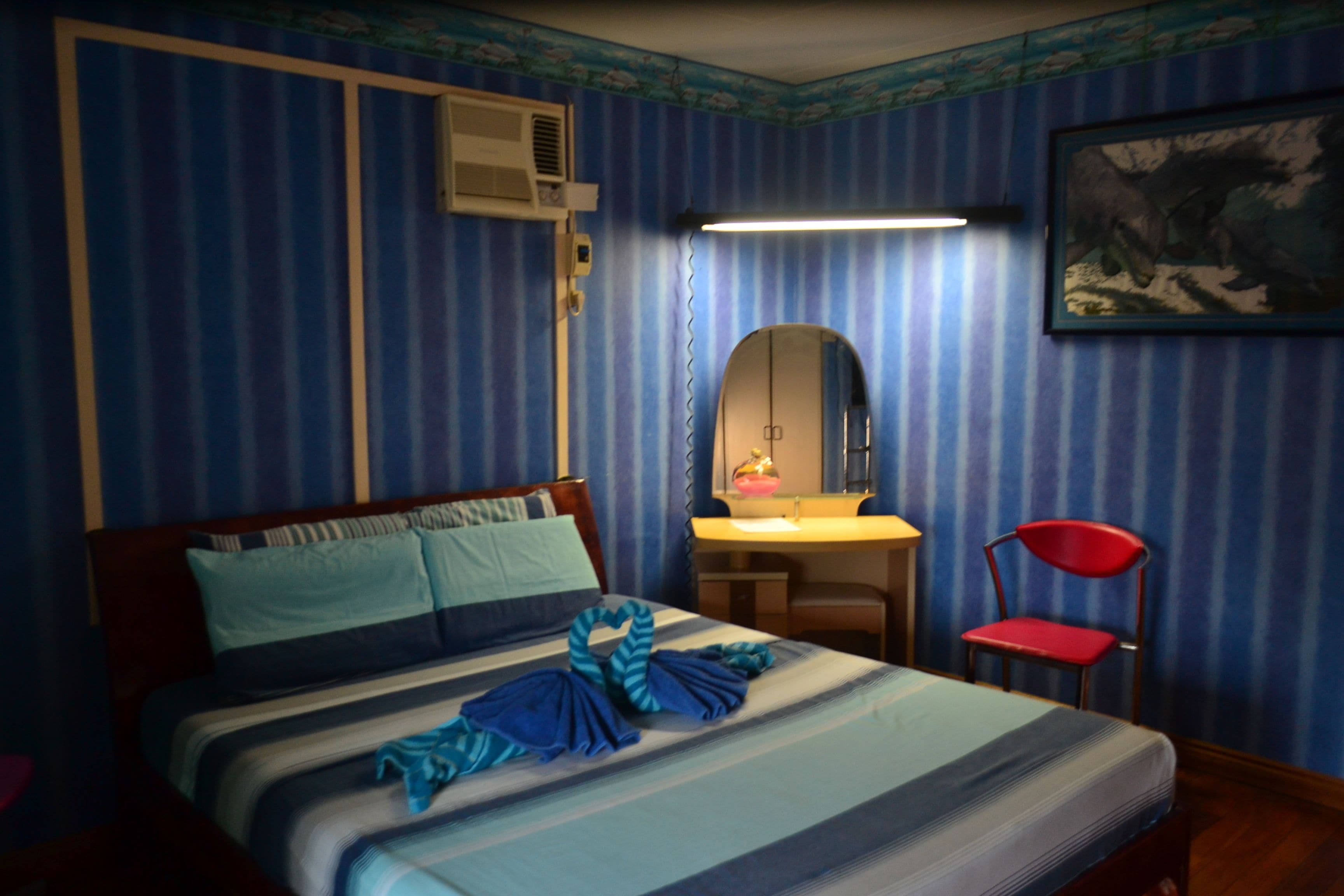 Une chambre d’hôtel avec un papier peint à rayures, un lit recouvert d’une courtepointe à rayures, un bureau avec un miroir, une chaise rouge et un climatiseur fixé au mur.