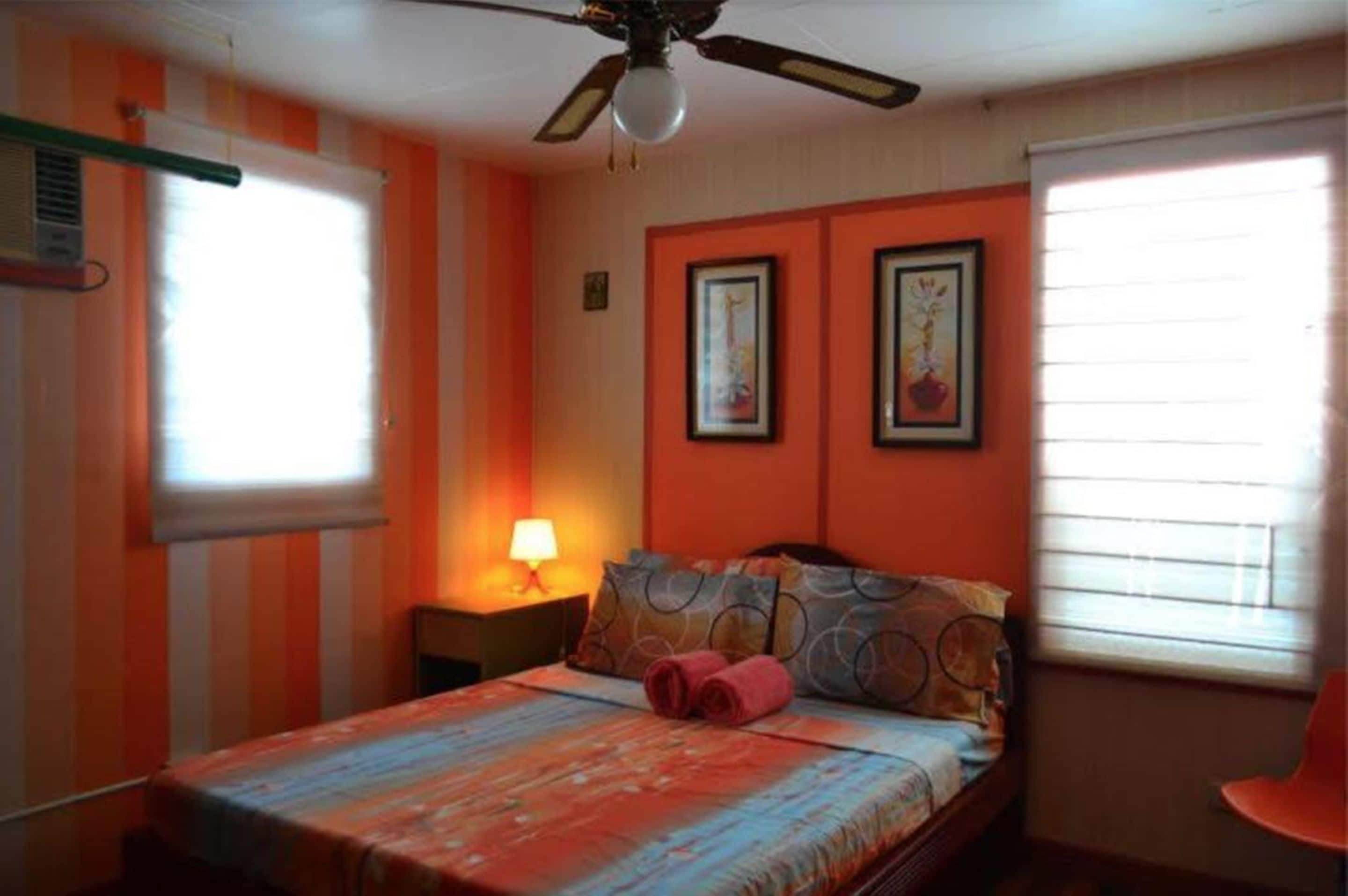 A bedroom with a bed, a ceiling fan, a window with blinds, and two framed pictures on the wall.