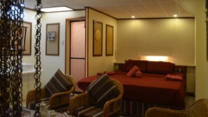 Une chambre d’hôtel avec un lit, deux fauteuils en osier, une table avec une théière et des tableaux encadrés accrochés au mur.