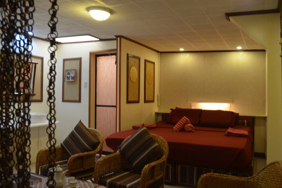 Una habitación de hotel con una cama, dos sillas de mimbre, una mesa con tetera y cuadros en la pared.