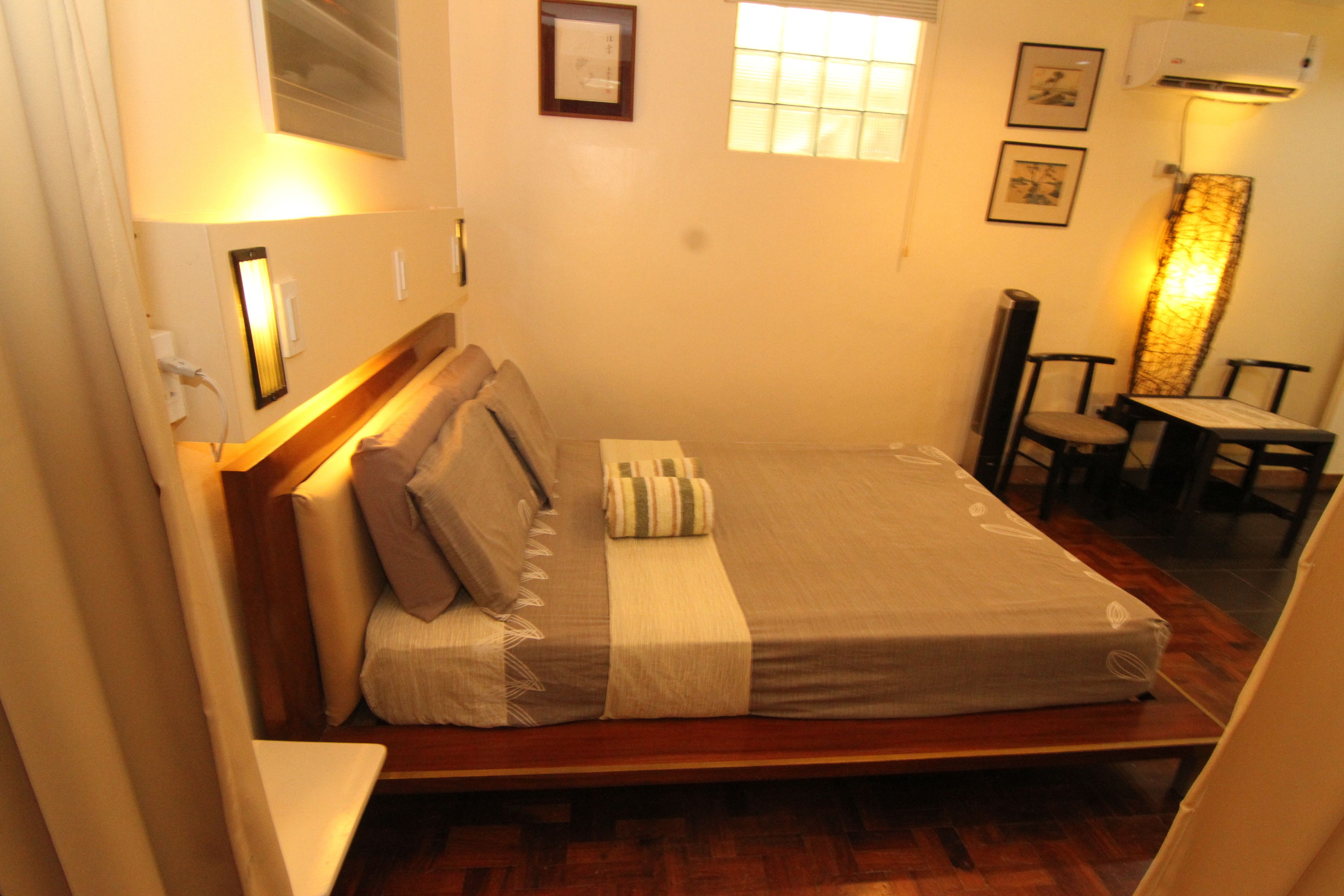 Una cama bien hecha con almohadas y una colcha, una mesita con sillas, y un aire acondicionado instalado en la pared.
