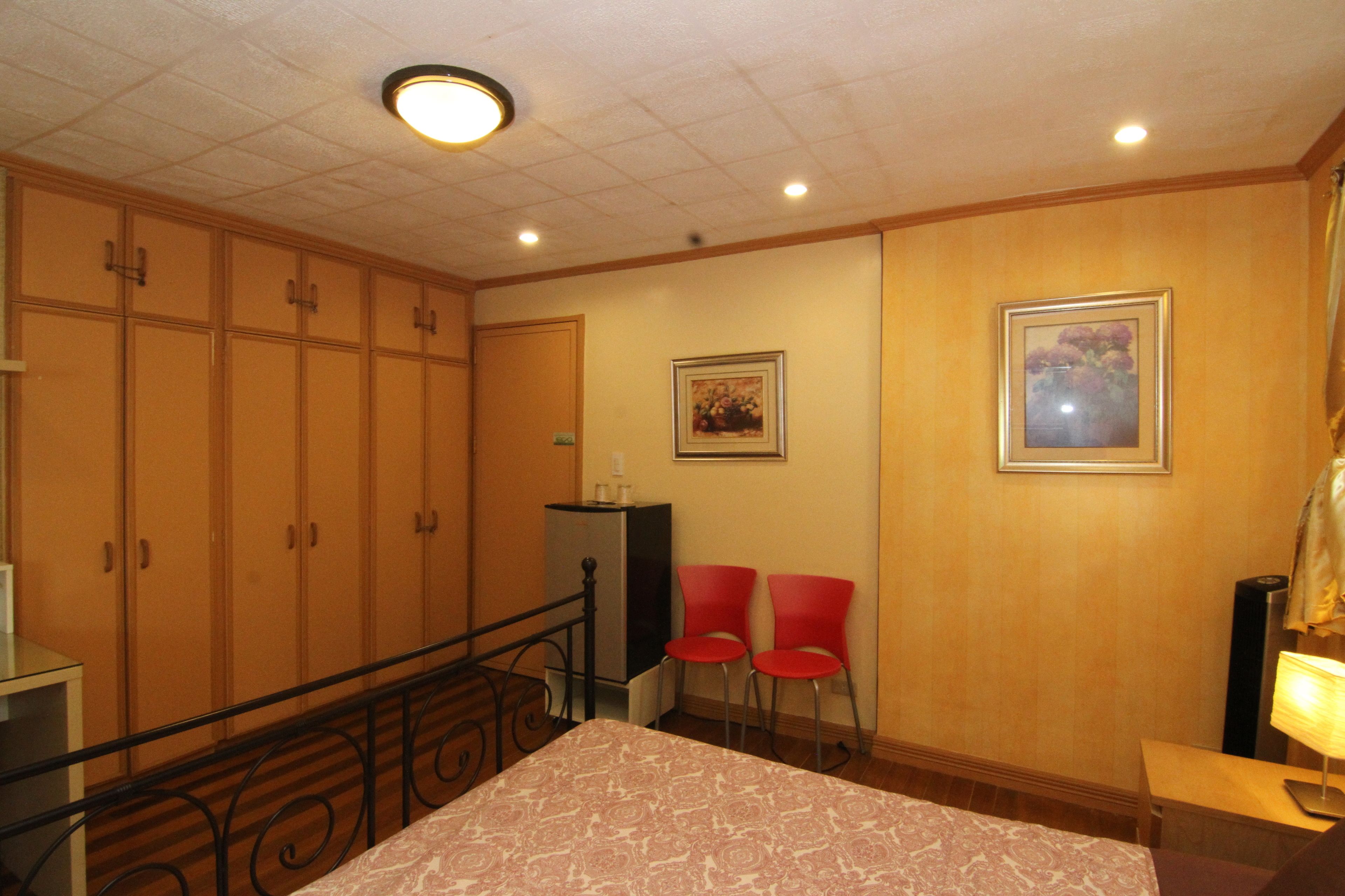 Una habitació d'hotel amb un llit, una taula de treball, dues cadires vermelles, una taula petita, un quadre a la paret i una làmpada al sostre.