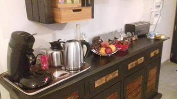 Un plan de travail de cuisine avec une machine à café, une bouilloire, une bouteille d’eau, un panier de fruits et un grille-pain.