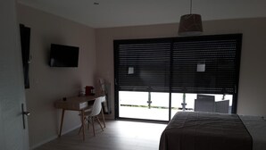 Un dormitorio con cama, escritorio y silla. Hay una ventana con persianas y un televisor montado en la pared.