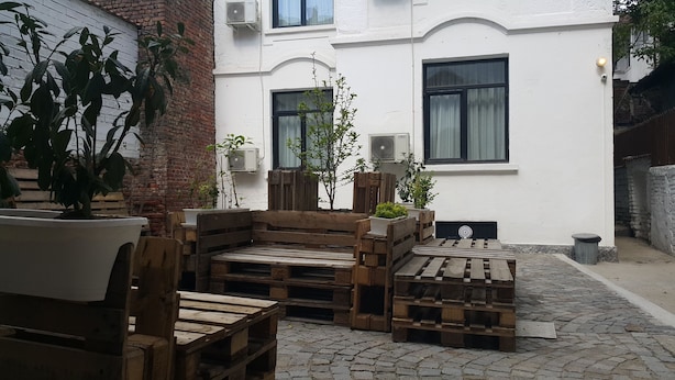A courtyard with wooden furniture and potted plants, a white building with multiple windows, and a brick wall.