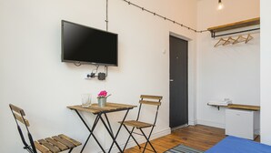 A compact room with a TV mounted on the wall, a small table with chairs, a mini-fridge, and a wooden bench.
