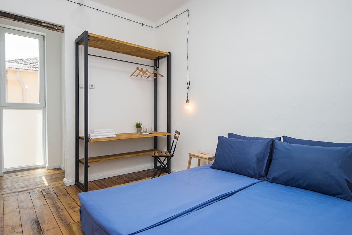 A bedroom with a bed, a wooden shelf with hanging clothes, a chair, and a small table.