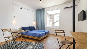 A compact bedroom with a bed, a small table, two chairs, a window with curtains, and a TV on the wall.