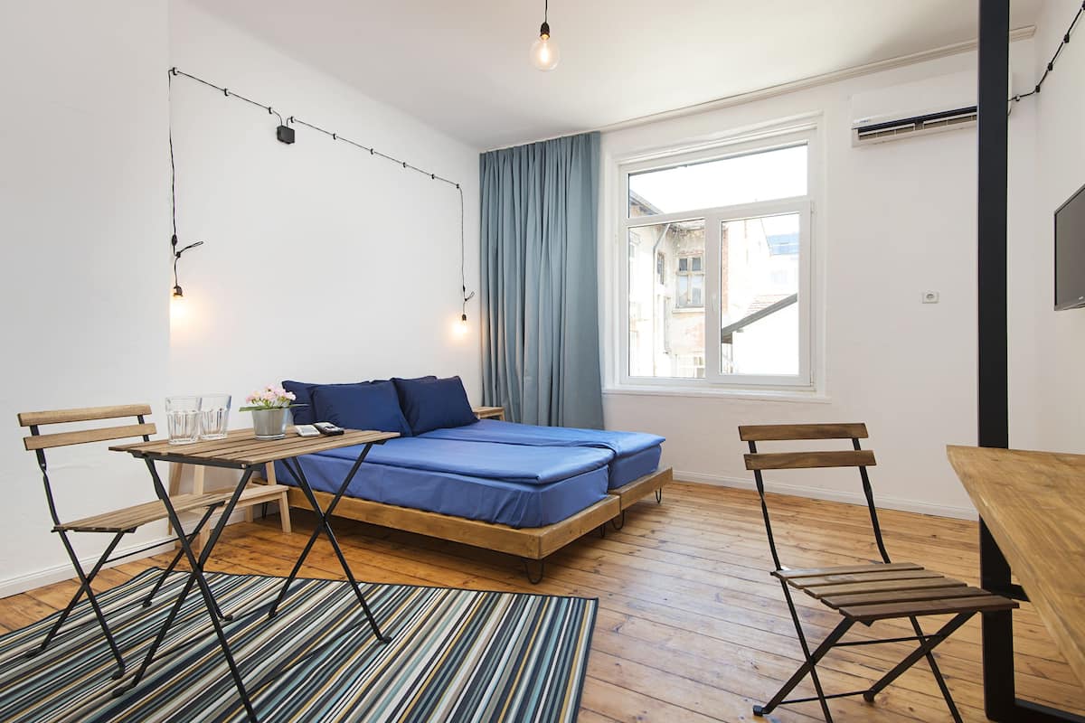 A compact bedroom with a bed, a small table, two chairs, a window with curtains, and a TV on the wall.