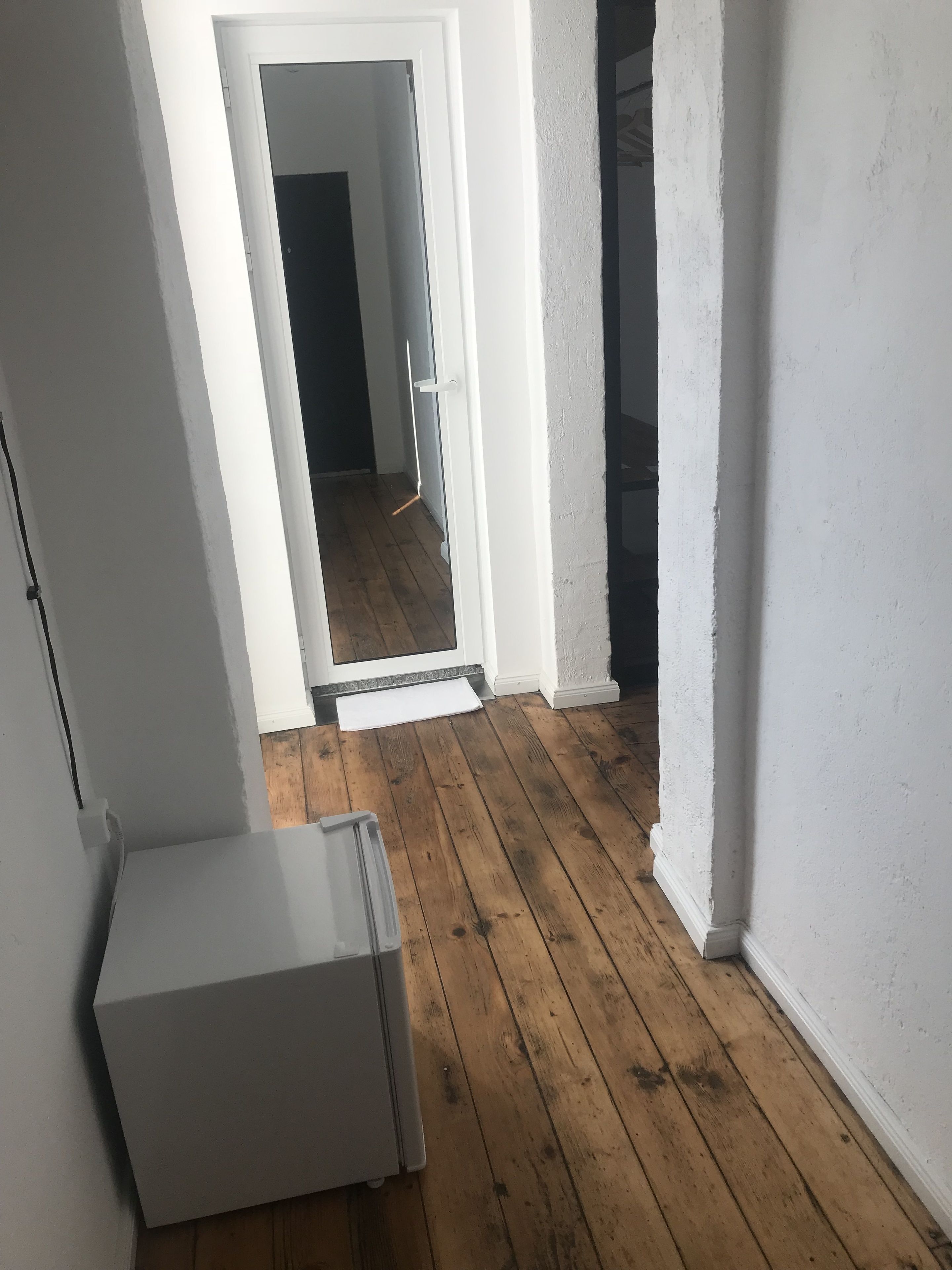 A narrow hallway with wooden flooring, white walls, and a white refrigerator in the foreground.