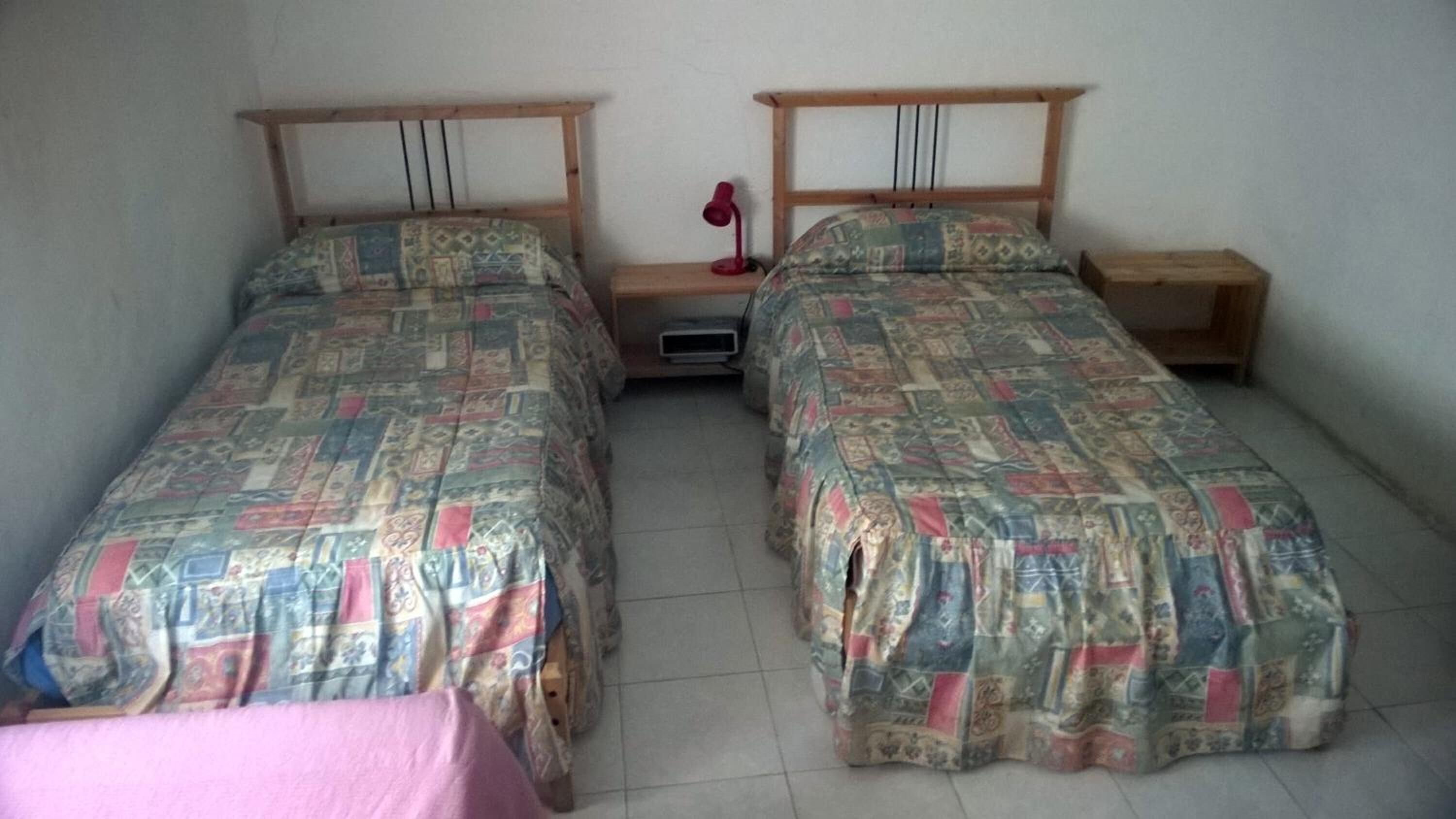 Dos camas individuales con cabeceros de madera, una mesita de noche con lámpara y suelo de baldosas.