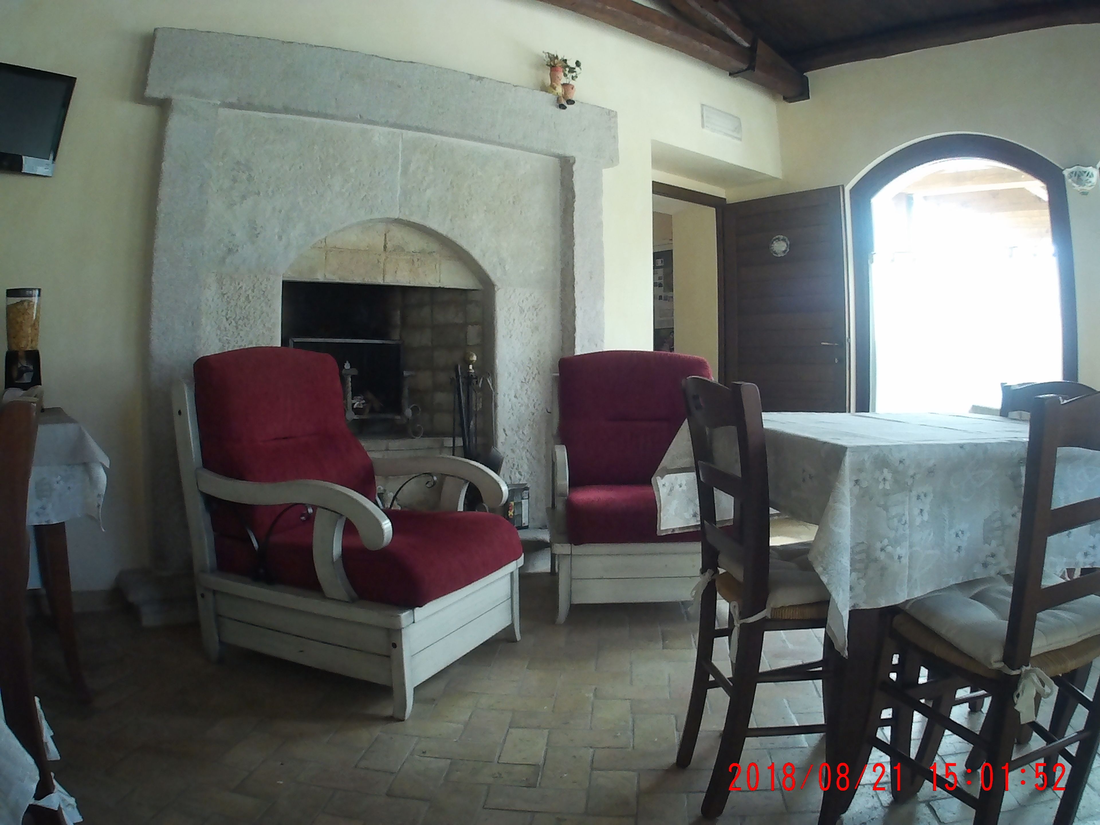 A cozy room with a fireplace, two red armchairs, a wooden table, and a view through an open door.