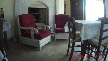 A cozy room with a fireplace, two red armchairs, a wooden table, and a view through an open door.