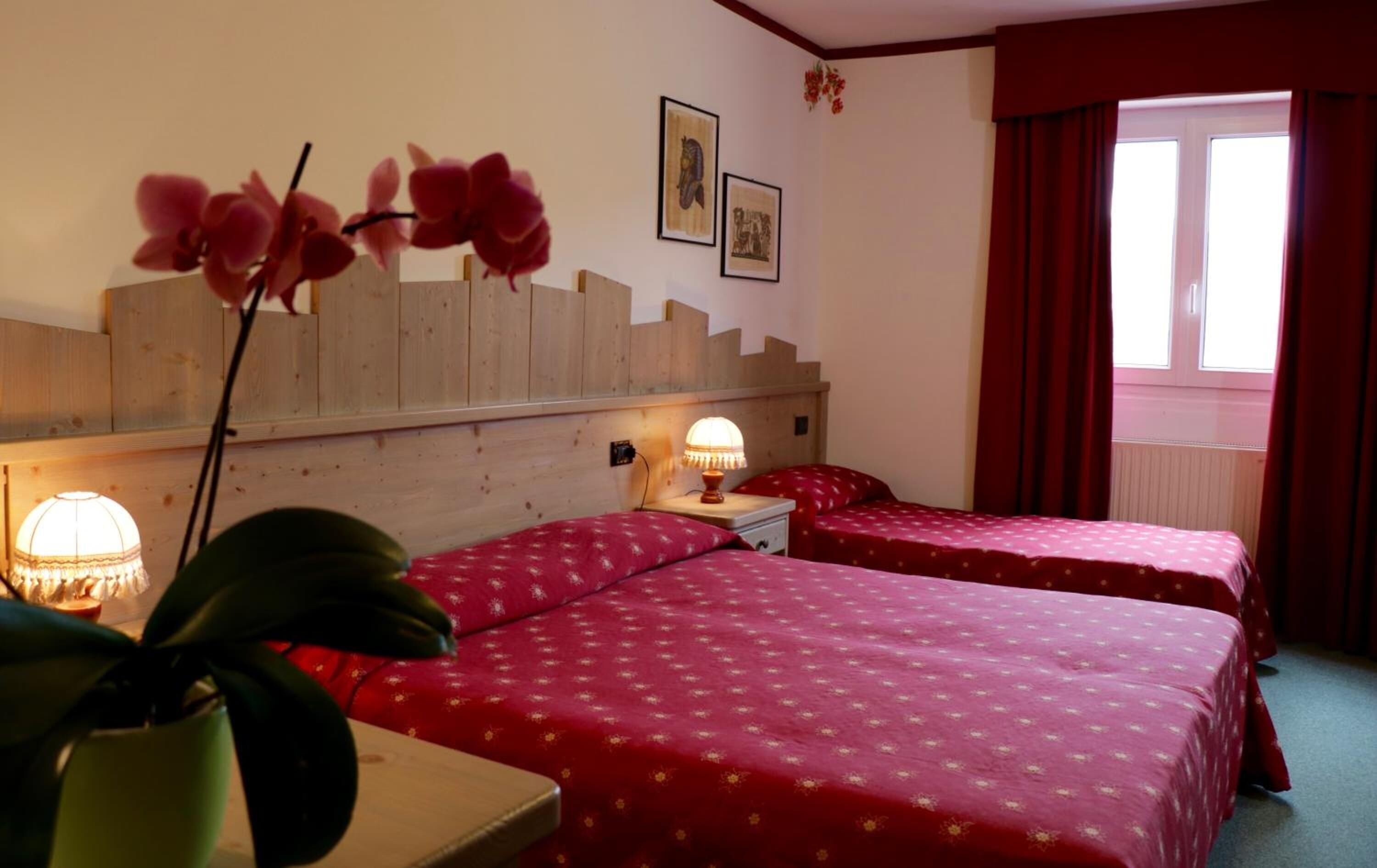 A hotel room with two beds, a wooden headboard, a nightstand with a lamp, and a window with red curtains.