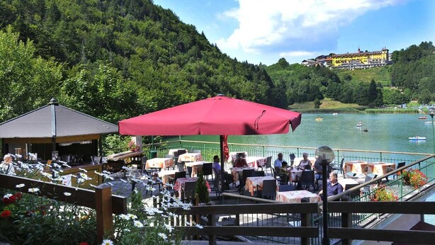 An outdoor dining area with tables and umbrellas, overlooking a lake with boats and a building in the background. - Hotel Al Lago (Folgaria)