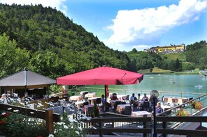 An outdoor dining area with tables and umbrellas, overlooking a lake with boats and a building in the background. - Hotel Al Lago (Folgaria)