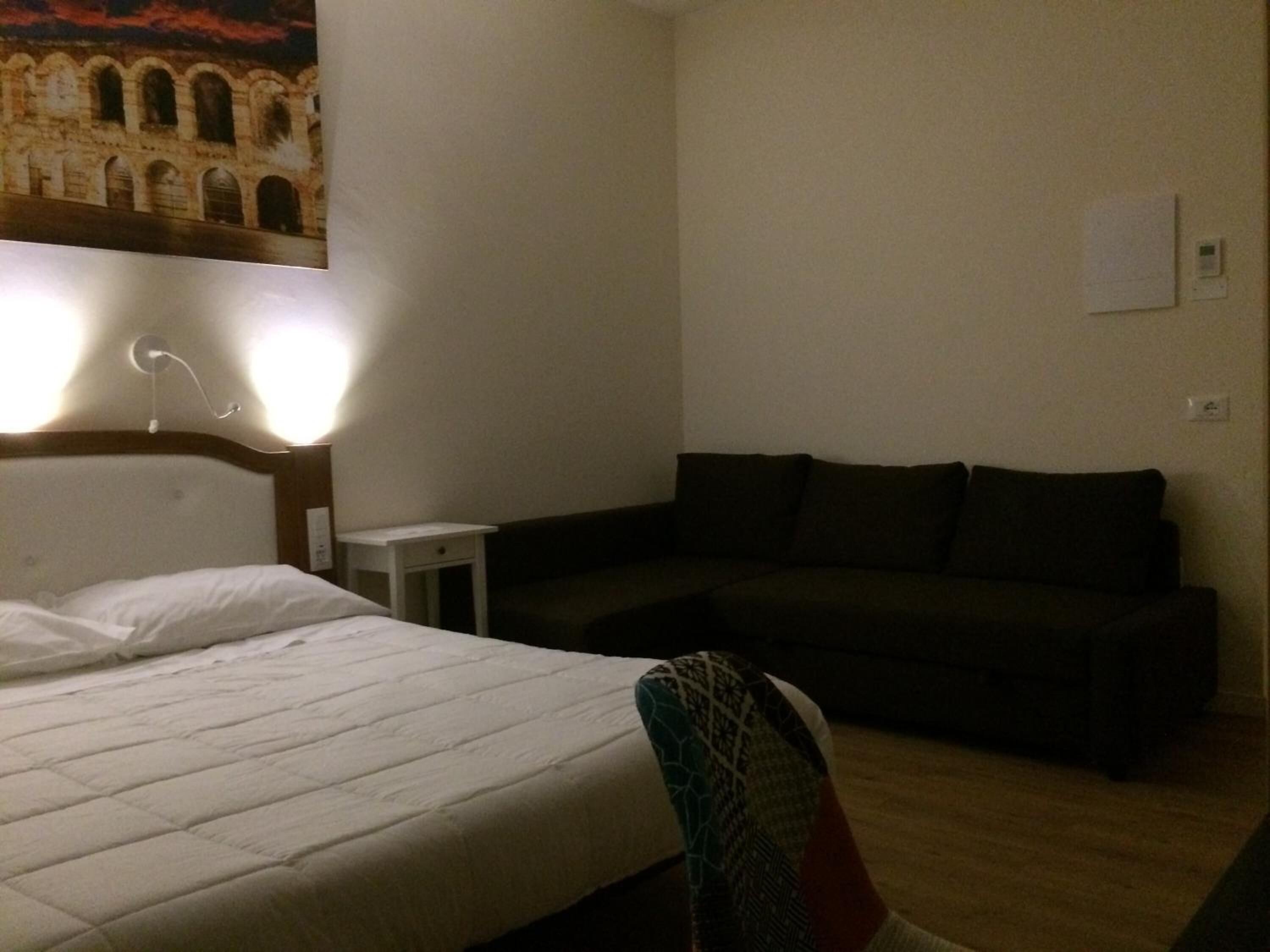 Habitación de hotel con cama, mesita de noche pequeña, sofá oscuro e iluminación empotrada en la pared.