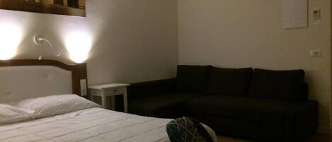 Habitación de hotel con cama, mesita de noche pequeña, sofá oscuro e iluminación empotrada en la pared.
