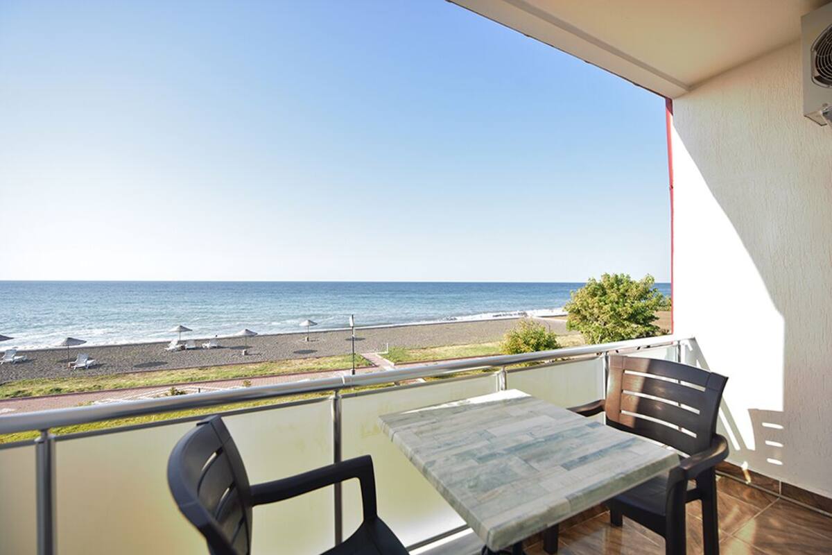 Un balcon avec une table et des chaises donnant sur une plage parsemée de parasols et bordée d’un sentier.