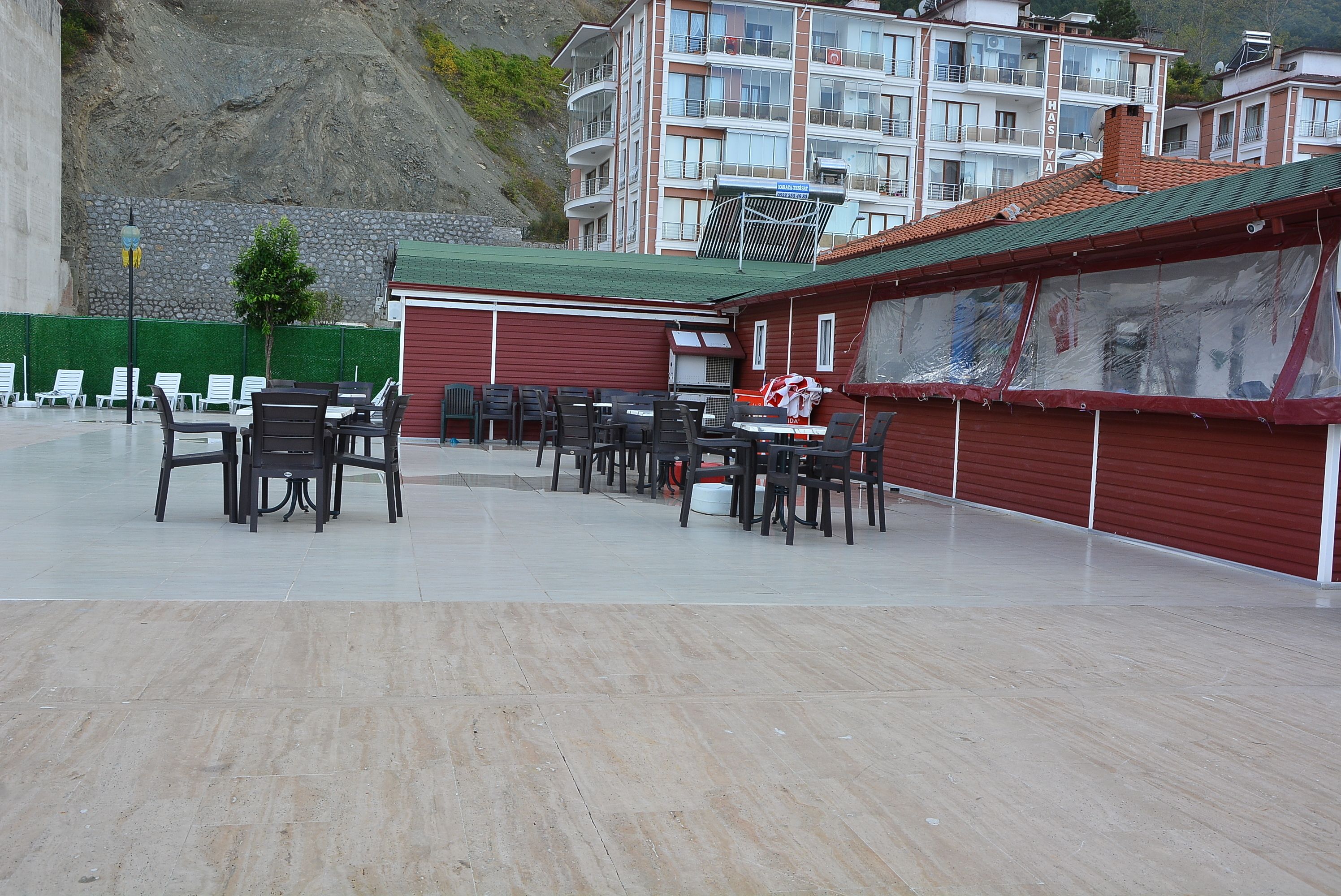 A covered outdoor seating area with tables and chairs, a red building with a green roof, and a stone wall in the background.