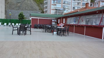 A covered outdoor seating area with tables and chairs, a red building with a green roof, and a stone wall in the background.