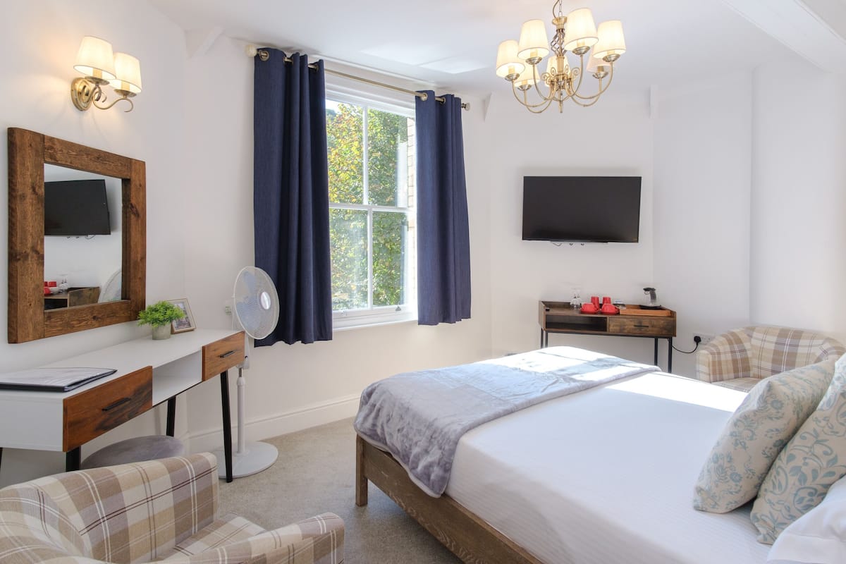 A bedroom with a bed, a desk, a chair, a TV, and a window with curtains.