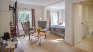 Een slaapkamer met een bed, twee fauteuils, een houten tafel met een bloevas, een televisie aan de muur en een raam met gordijnen.