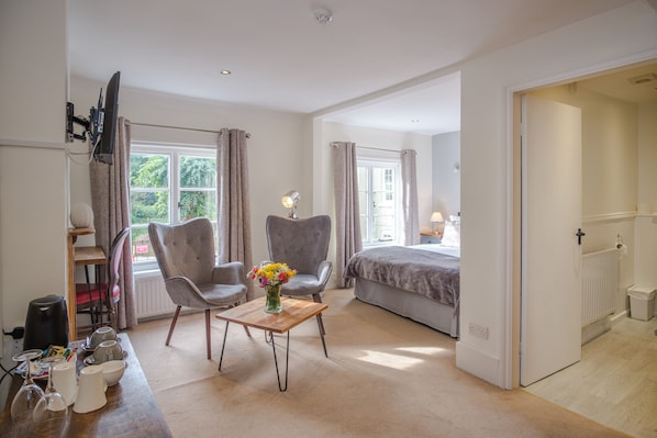 A bedroom with a bed, two armchairs, a wooden table with a vase of flowers, a television mounted on the wall, and a window with curtains.
