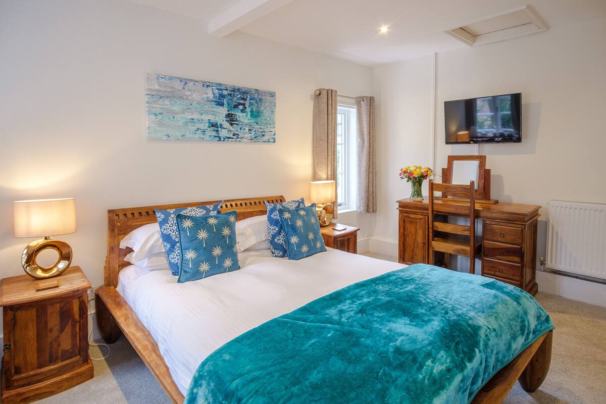 A bedroom with a bed, wooden nightstands, a TV, a painting, and a window with curtains.