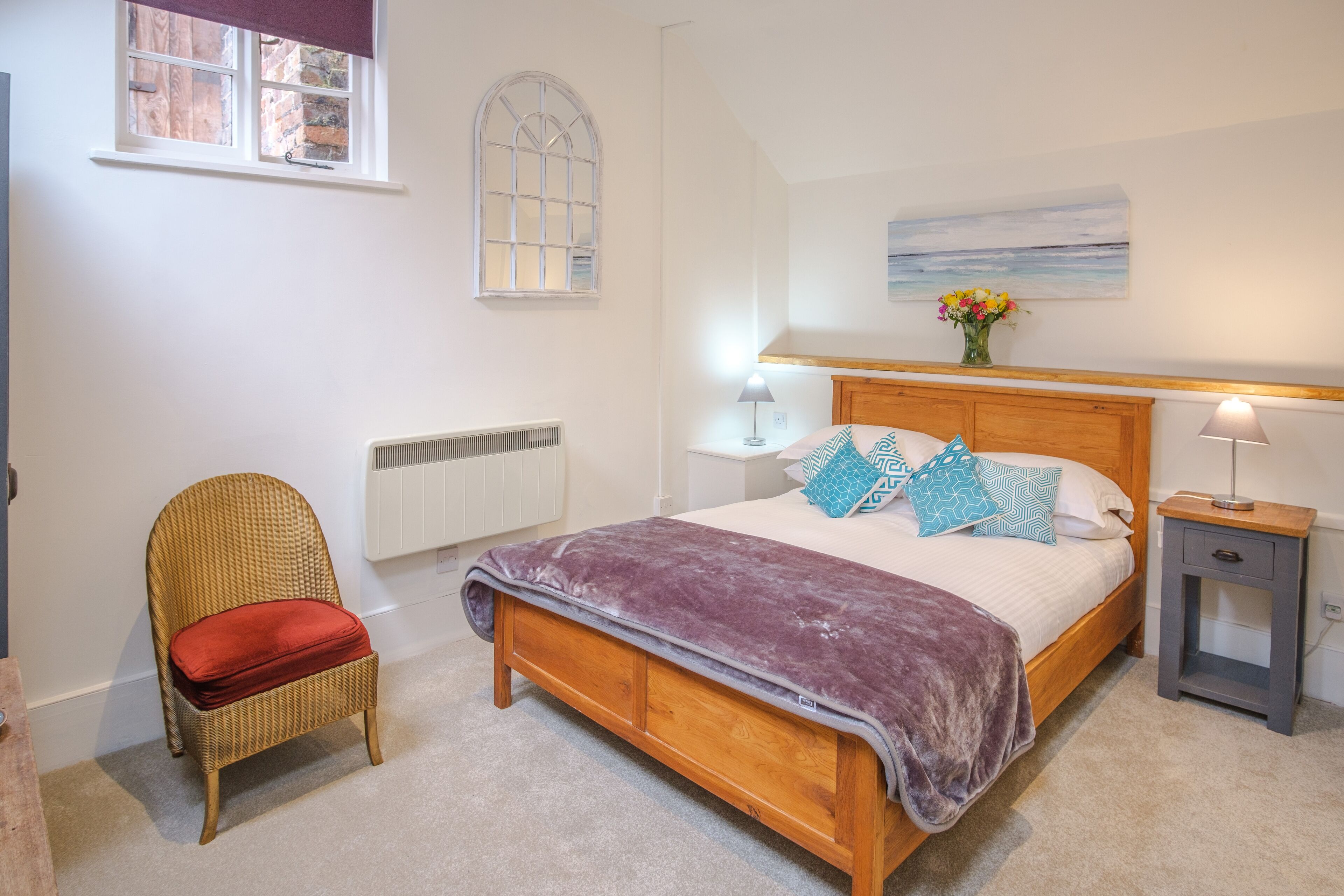 Een slaapkamer met een houten bed, een rieten stoel met een rode kussen, een radiator, een raam en een nachtkastje met een lamp.