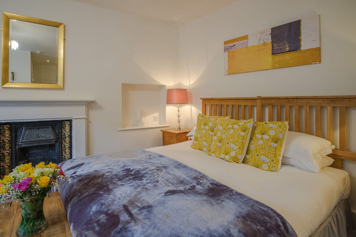 A bedroom with a bed, a mirror, a fireplace, and a wall-mounted artwork.