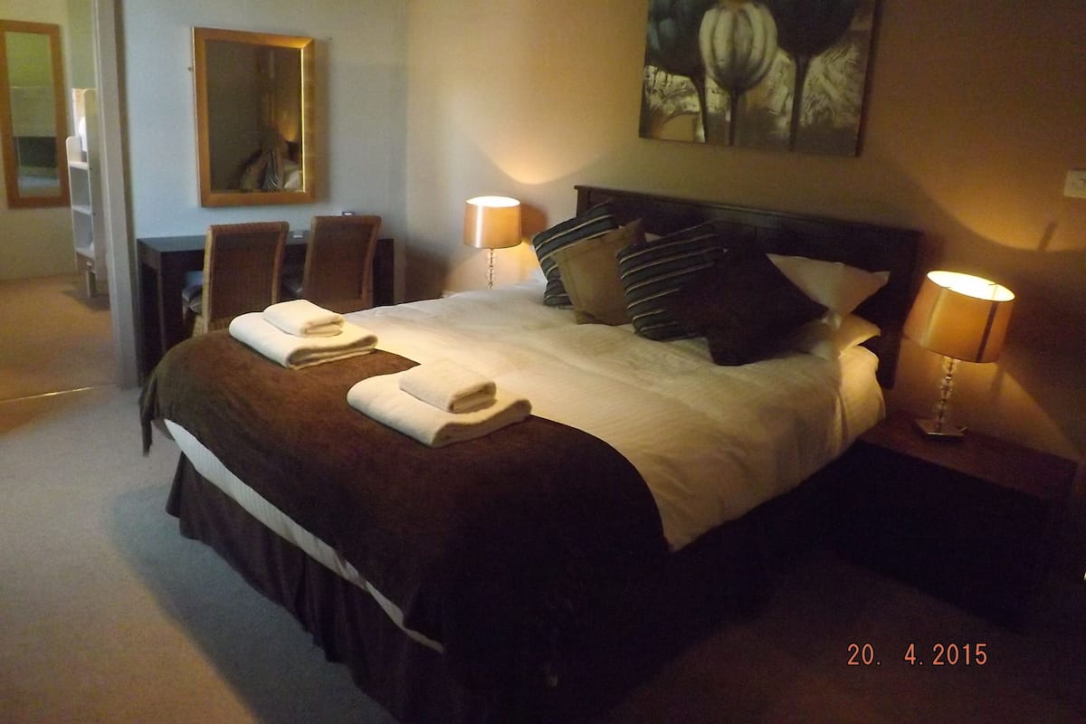 A hotel room with a large bed, two bedside lamps, a painting on the wall, and a desk with a chair.