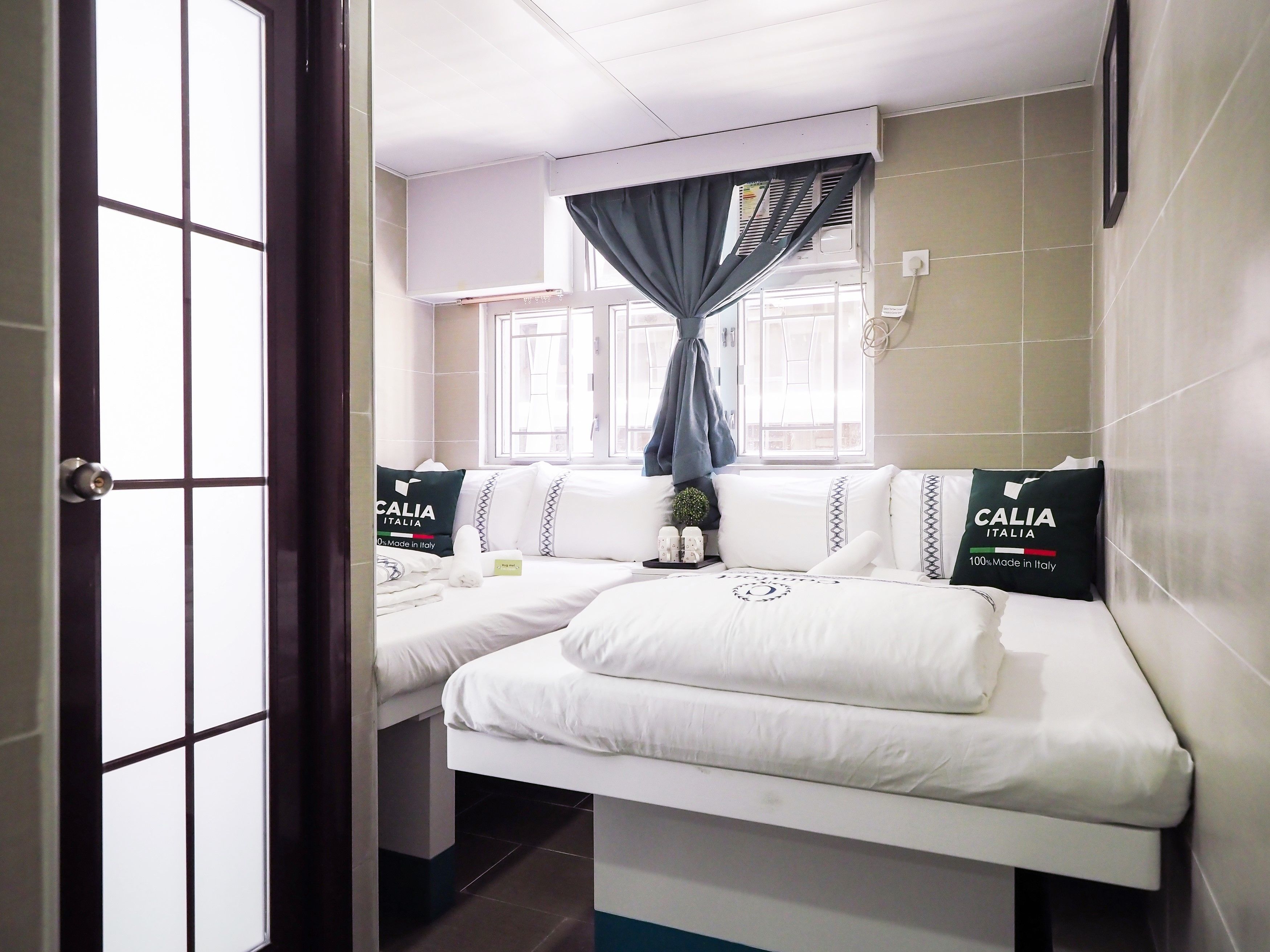 A hotel room with two beds, a window with curtains, and a wall-mounted air conditioner.