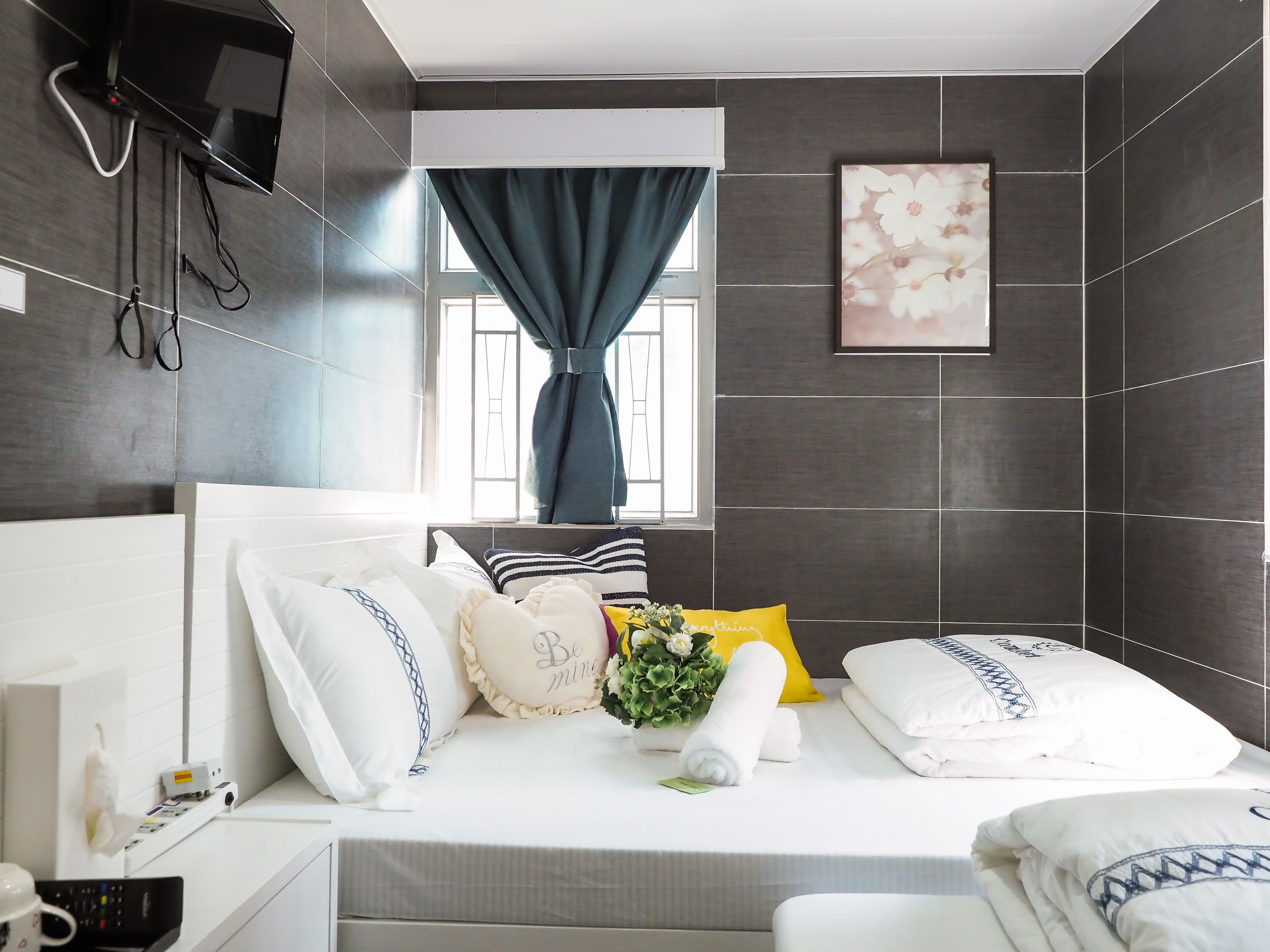 A modern hotel room with a neatly made bed, a window with curtains, a flat-screen TV mounted on the wall, and a small bedside table with a remote control.