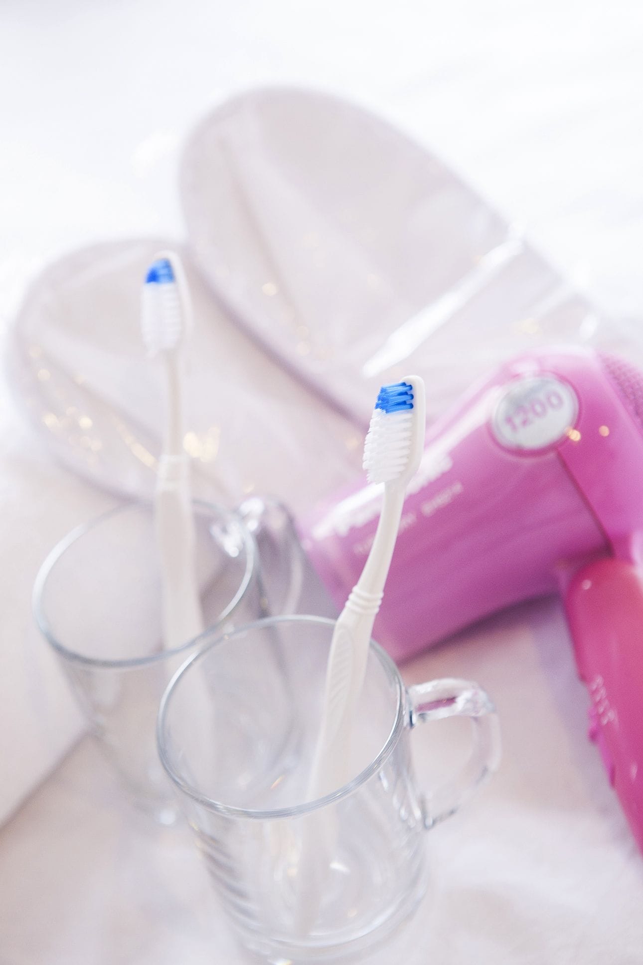 Two toothbrushes in clear glass cups, a pink toothbrush case, and a pair of white slippers.