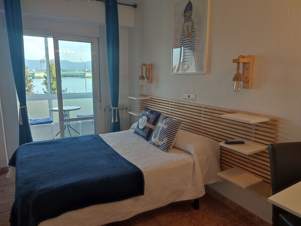 Un dormitorio con cama, escritorio, silla, vistas al exterior y un cuadro en la pared.