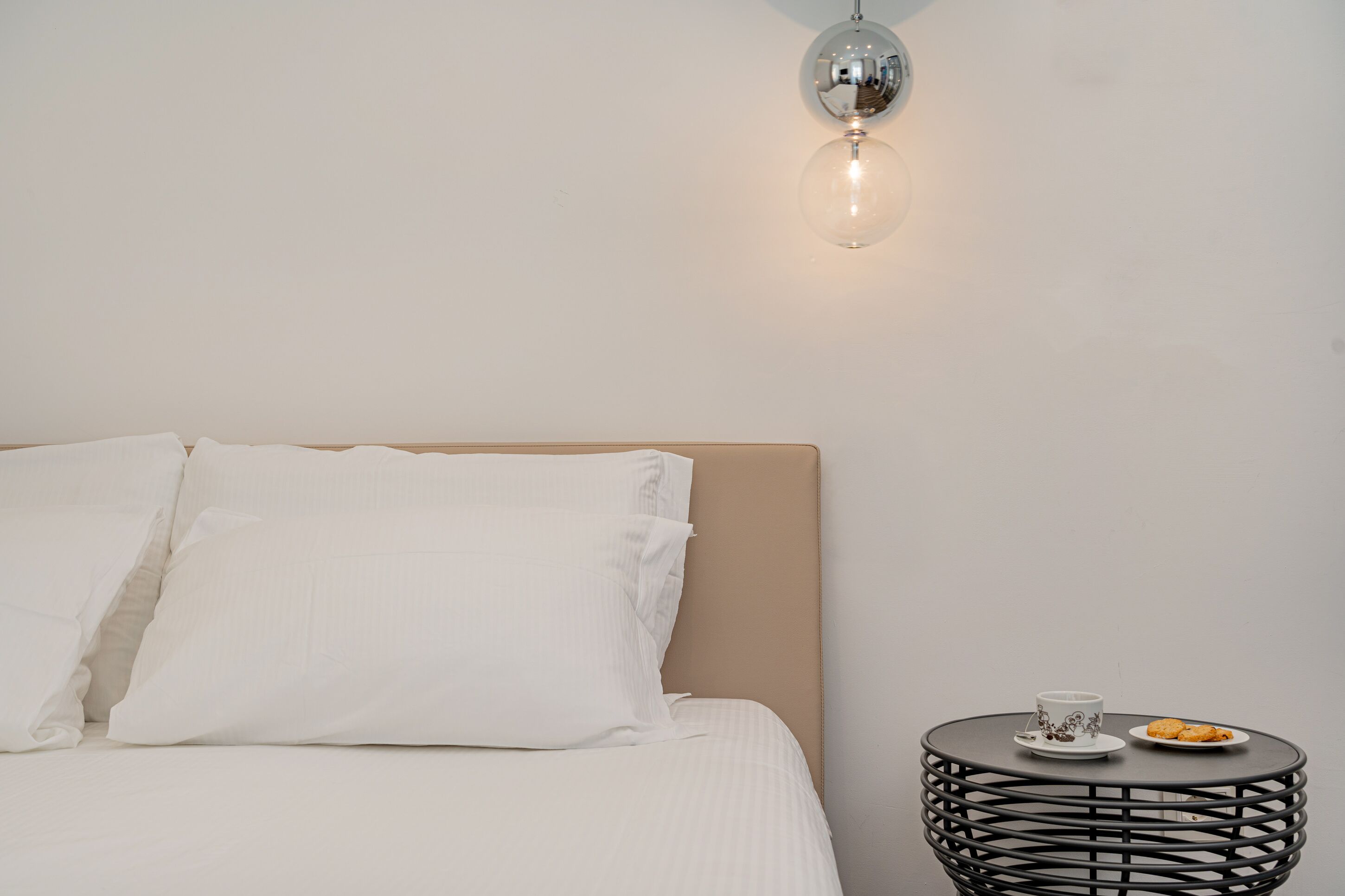 Ein ordentlich bezogenes Bett mit weißen Leinen, ein runder schwarzer Nachttisch mit einer Tasse und Keksen sowie eine wandmontierte Lampe.