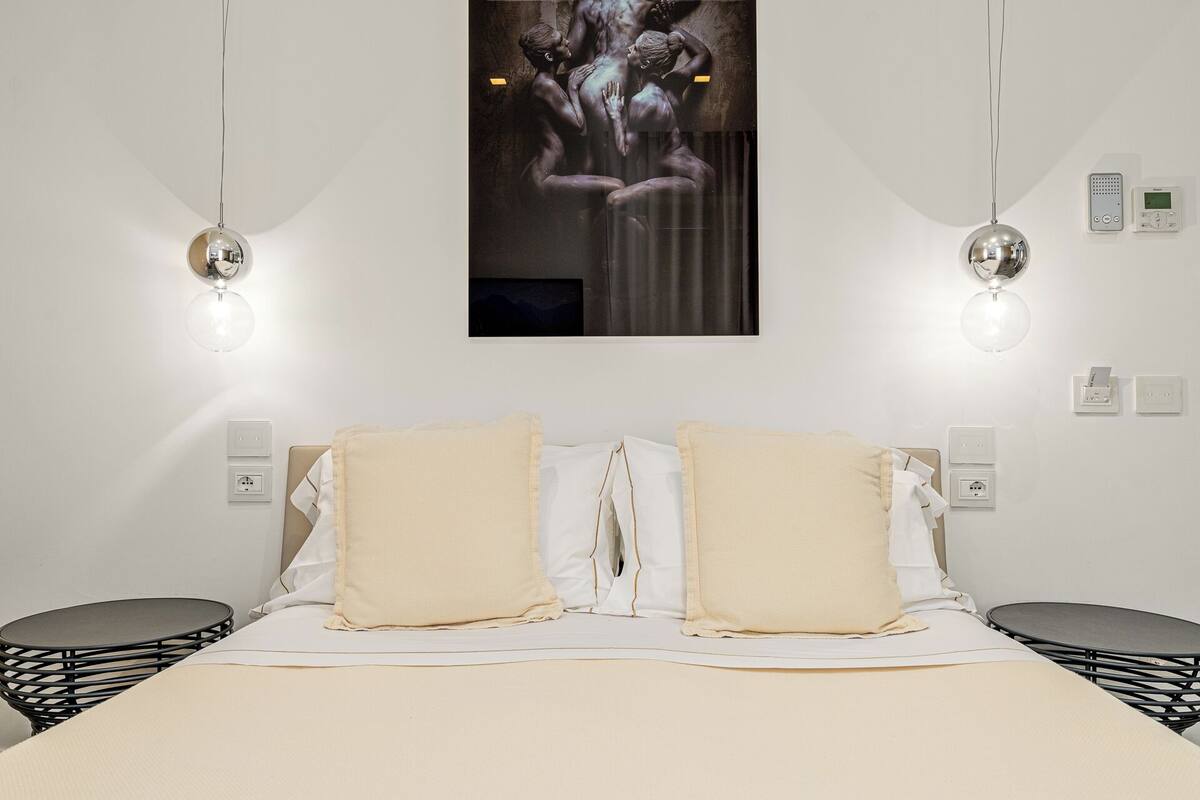 Un dormitorio moderno con una cama grande, dos mesitas de noche, una lámpara fijada a la pared y una obra de arte enmarcada sobre la cama.