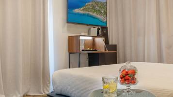 Une chambre d’hôtel équipée d’un lit, d’un téléviseur fixé au mur, d’un bureau avec un ordinateur et d’une table sur laquelle se trouvent un verre d’eau et une corbeille de fruits.