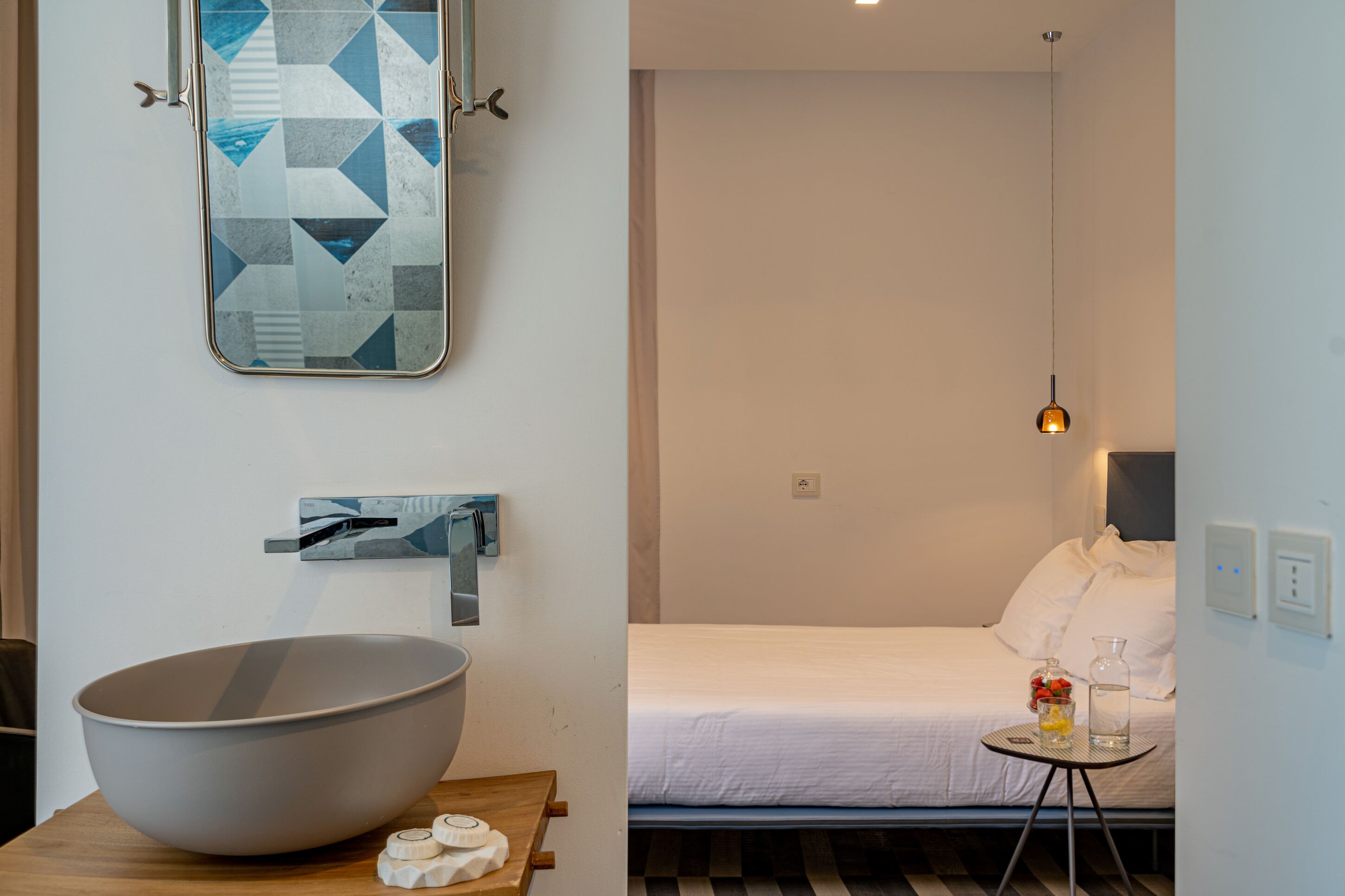 Una habitación de hotel moderna con un tazón grande sobre una mesa de madera, una cama con ropa blanca, una pequeña mesa redonda con un florero y una botella, y una obra de arte mural geométrica.