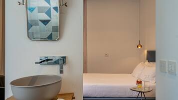 Una habitación de hotel moderna con un tazón grande sobre una mesa de madera, una cama con ropa blanca, una pequeña mesa redonda con un florero y una botella, y una obra de arte mural geométrica.