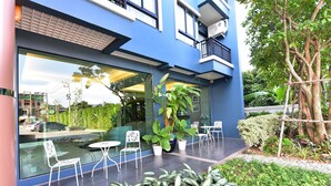 A modern building with large glass windows, outdoor seating area with white chairs and tables, and a well-maintained garden with various plants. - The U-Time Hotel (Hat Yai)