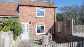 A brick house with two windows, a door, and a small patio with a chair and table.