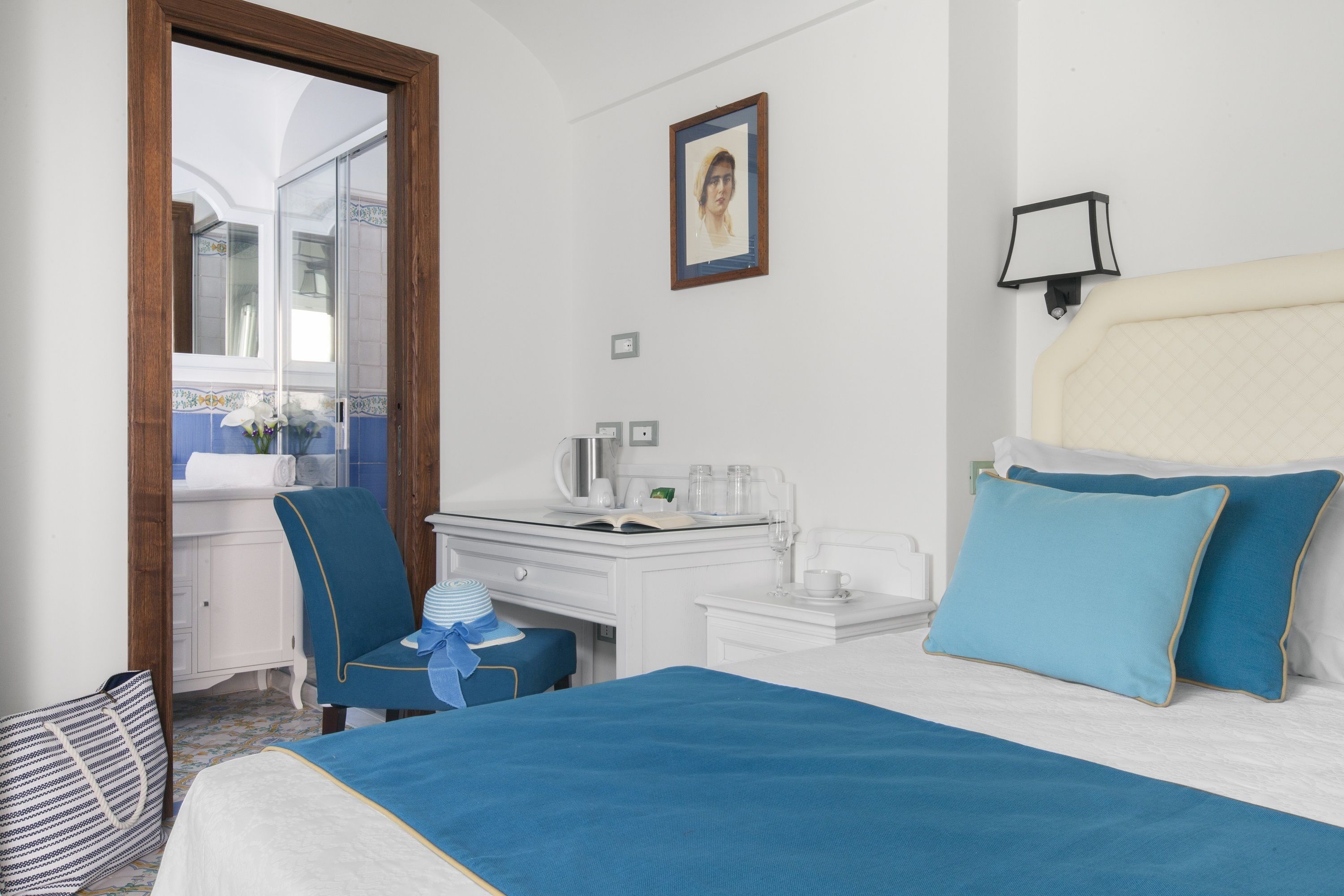 A hotel room with a bed, a desk, a chair, a small table, a mirror, and a picture on the wall.